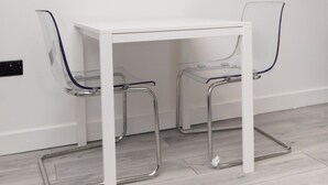 A white table with two clear chairs, a mounted TV displaying 'NETFLIX', and a white wall. - The Oasis Apartment (Leeds)