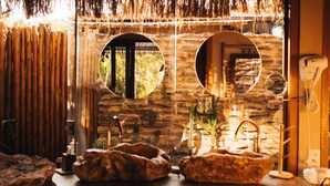 Une salle de bain rustique avec un mur en pierre, deux miroirs ronds, une table en bois avec un lavabo et un téléphone.