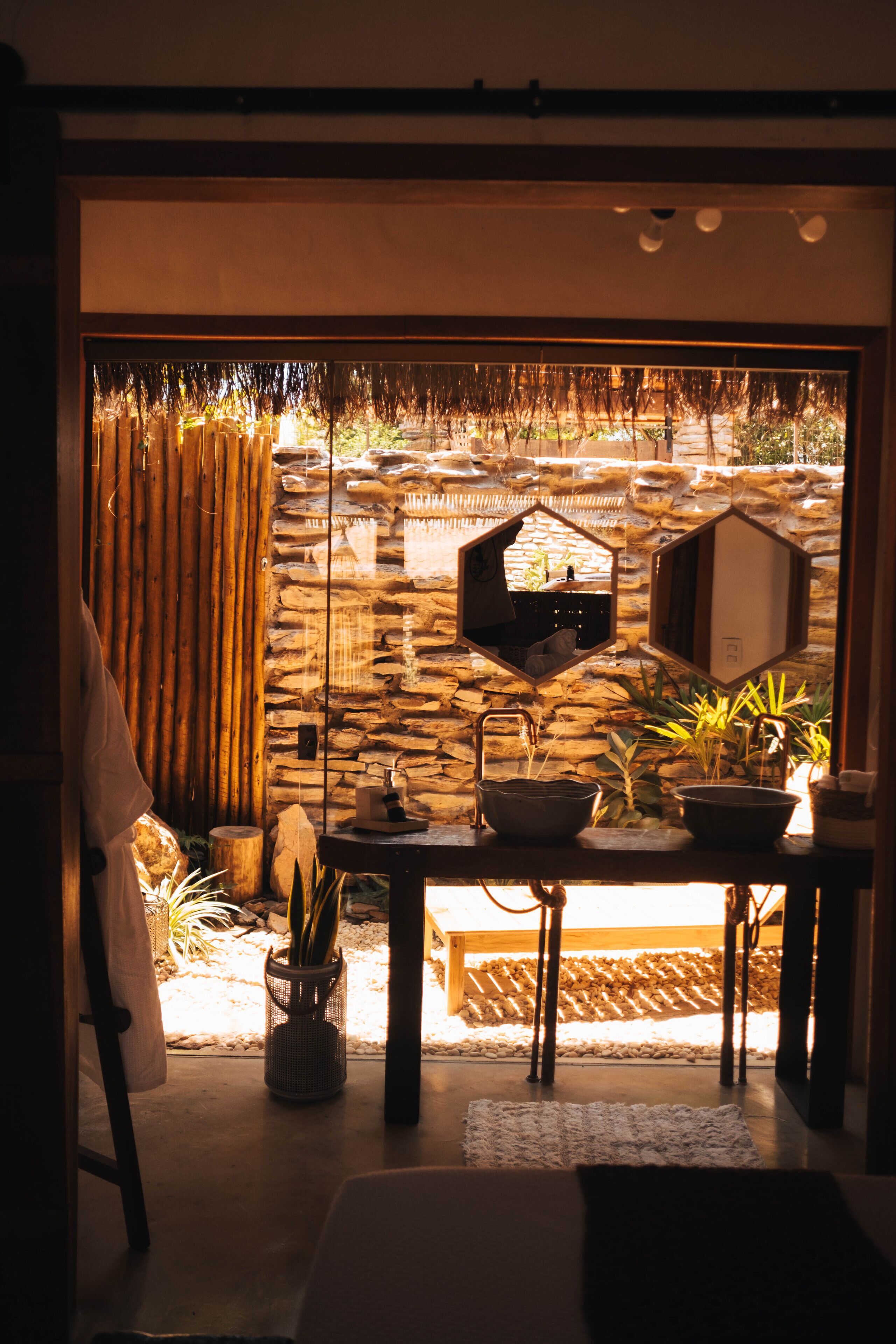 A room with a stone wall, a wooden table with a bowl and a potted plant, and a hanging mirror.