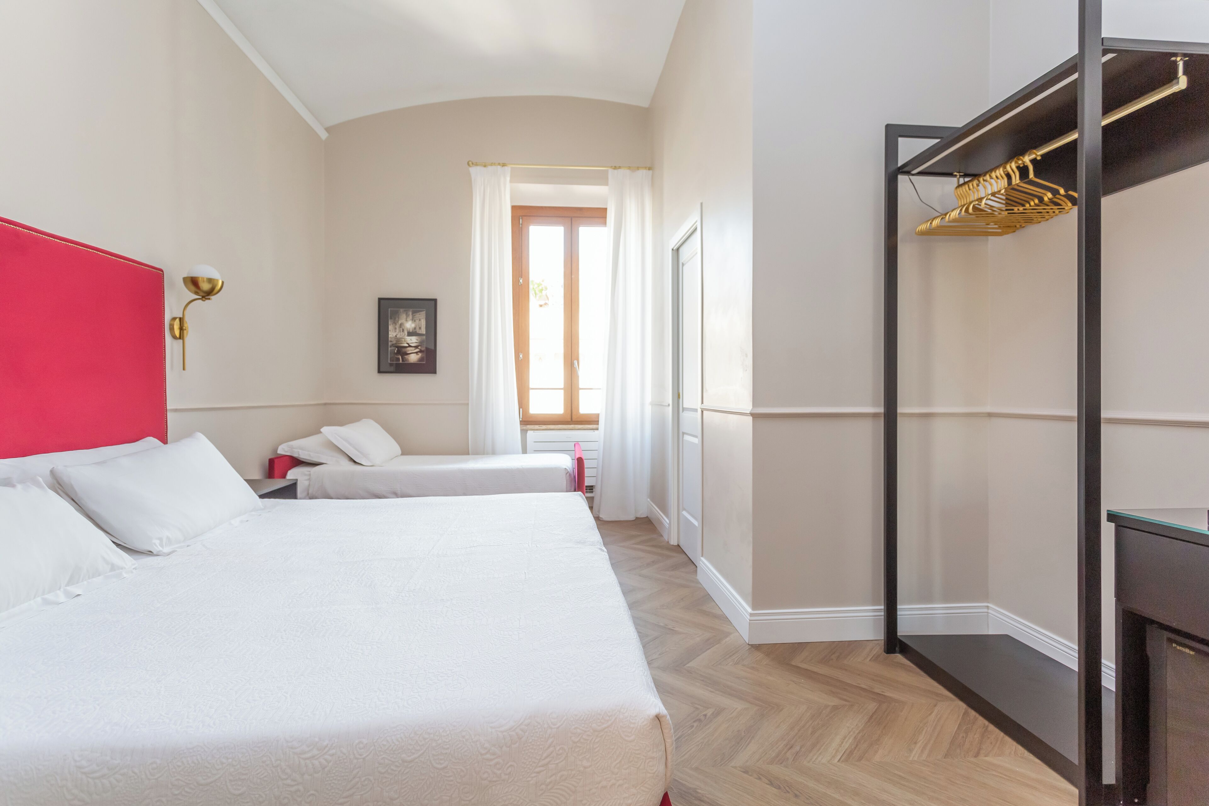 Ein Hotelzimmer mit zwei Betten, einem roten Kopfteil, einem Schrank mit Spiegel und einem Fenster mit Vorhängen.