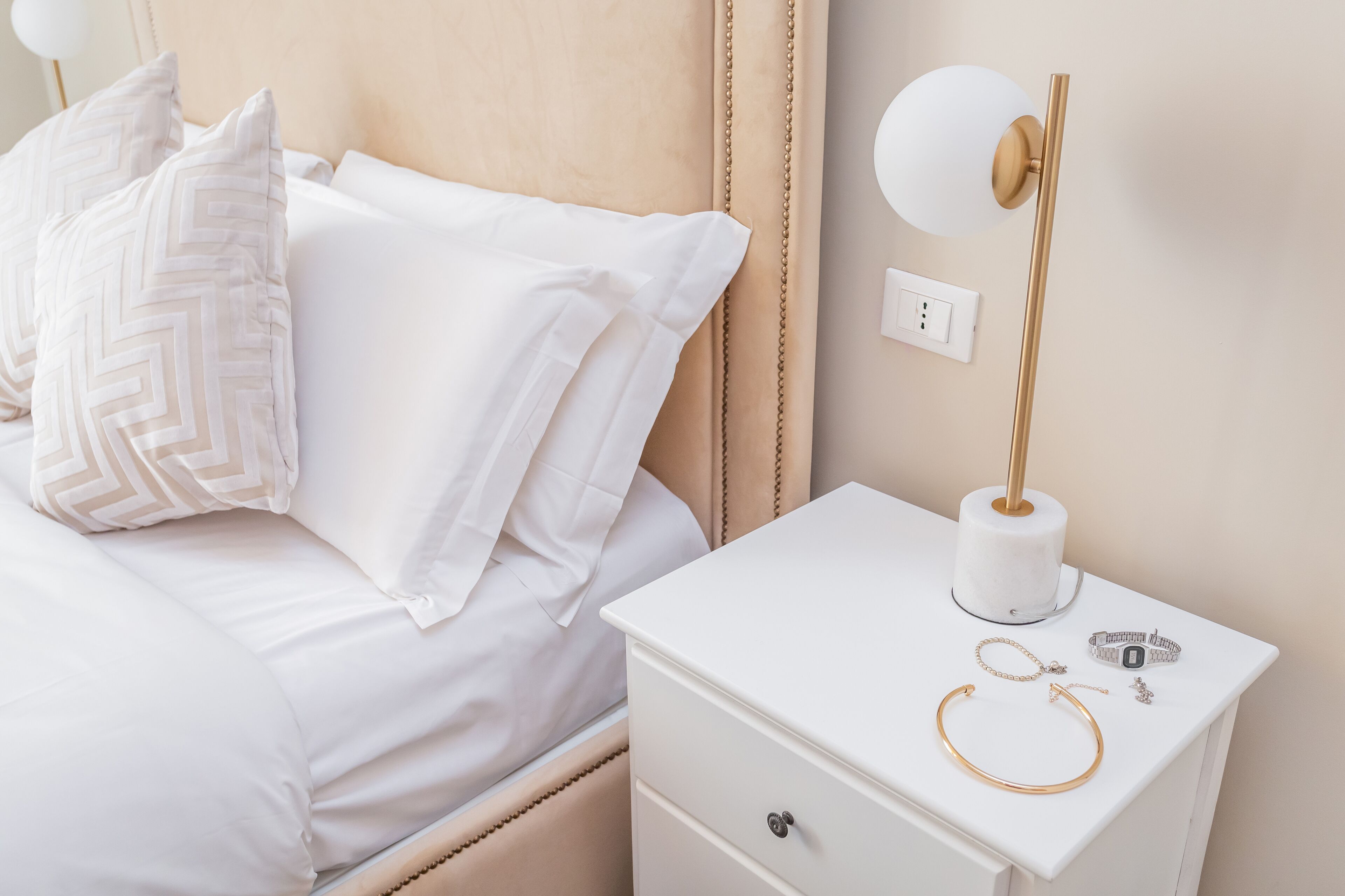 A neatly made bed with white bedding and decorative pillows, a bedside table with a lamp, and a small drawer.