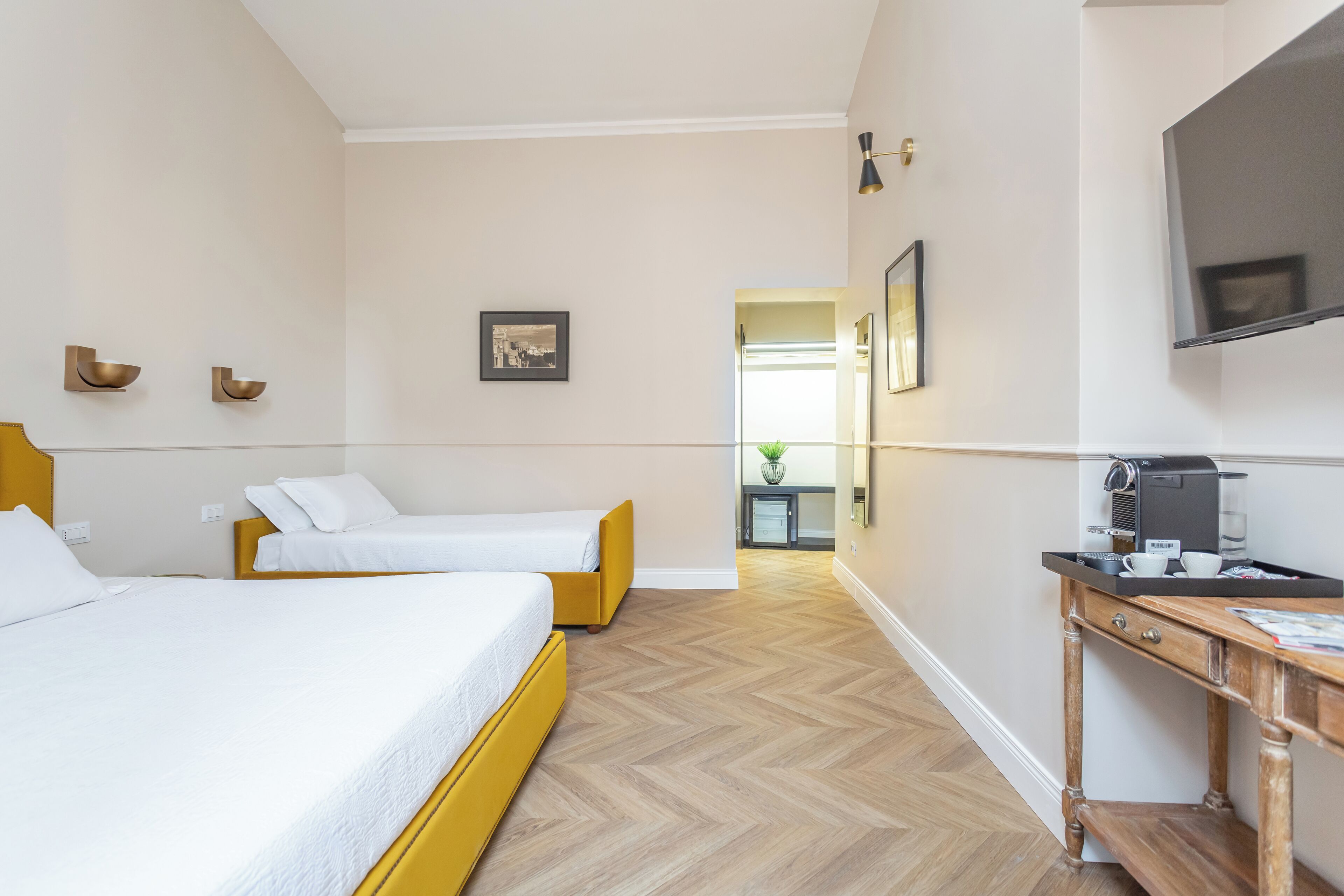 Habitación de hotel con dos camas, piso de madera, escritorio con cafetera y televisor en la pared.