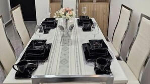 Ein Esszimmer mit einem langen Tisch, gedeckt für sechs Personen, umgeben von Stühlen. Es gibt eine Holzregal-Einheit mit Dekoration und einem Blumentopf.