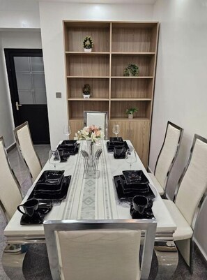 Un comedor con una larga mesa dispuesta para seis personas, rodeada de sillas. Se observa un aparador de madera con objetos decorativos y una planta en maceta.
