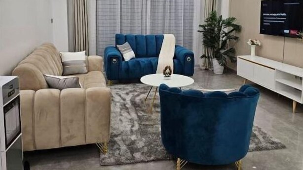 Ein modernes Wohnzimmer mit einem beigefarbenen Sofa, einem blauen Sofa, einem blauen Sessel und einem weißen Couchtisch. Ein Fernseher ist an der Wand montiert und eine Deckenleuchte ist vorhanden.