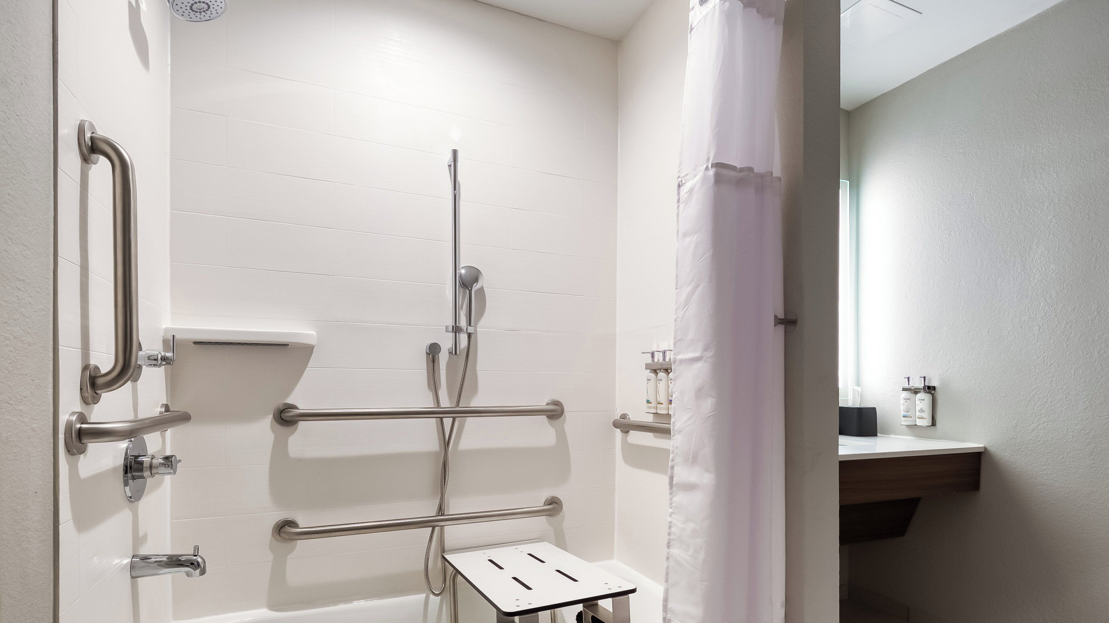 A bathroom with white tiled walls, a shower with a handheld showerhead, grab bars, and a small step stool.
