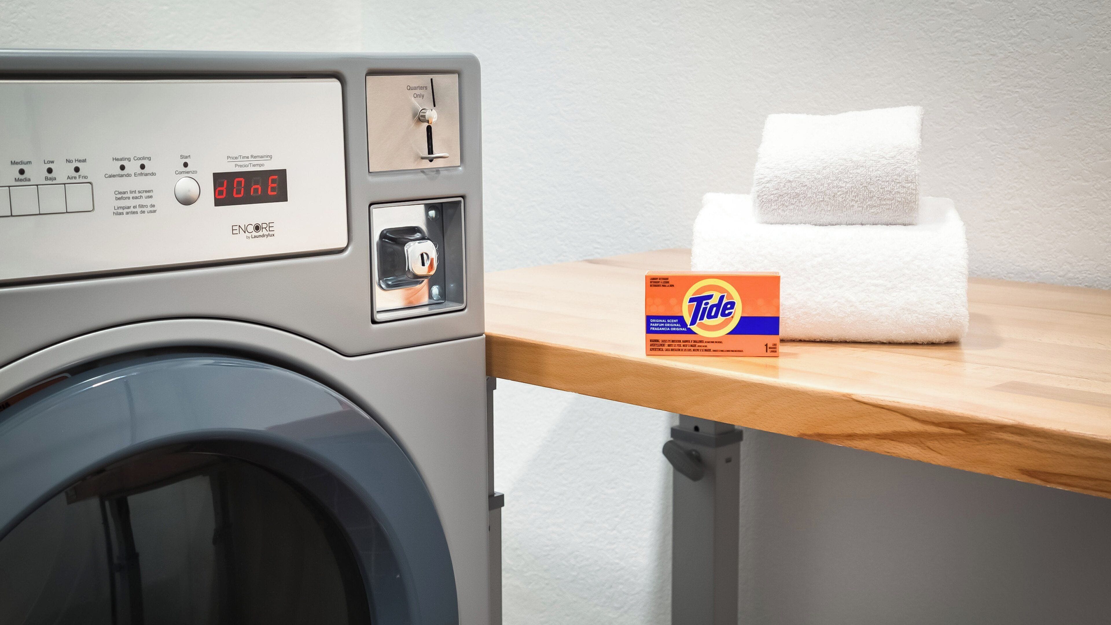 A front-loading washing machine with a control panel, a timer set to 0:00, and a Tide detergent box on a wooden table.