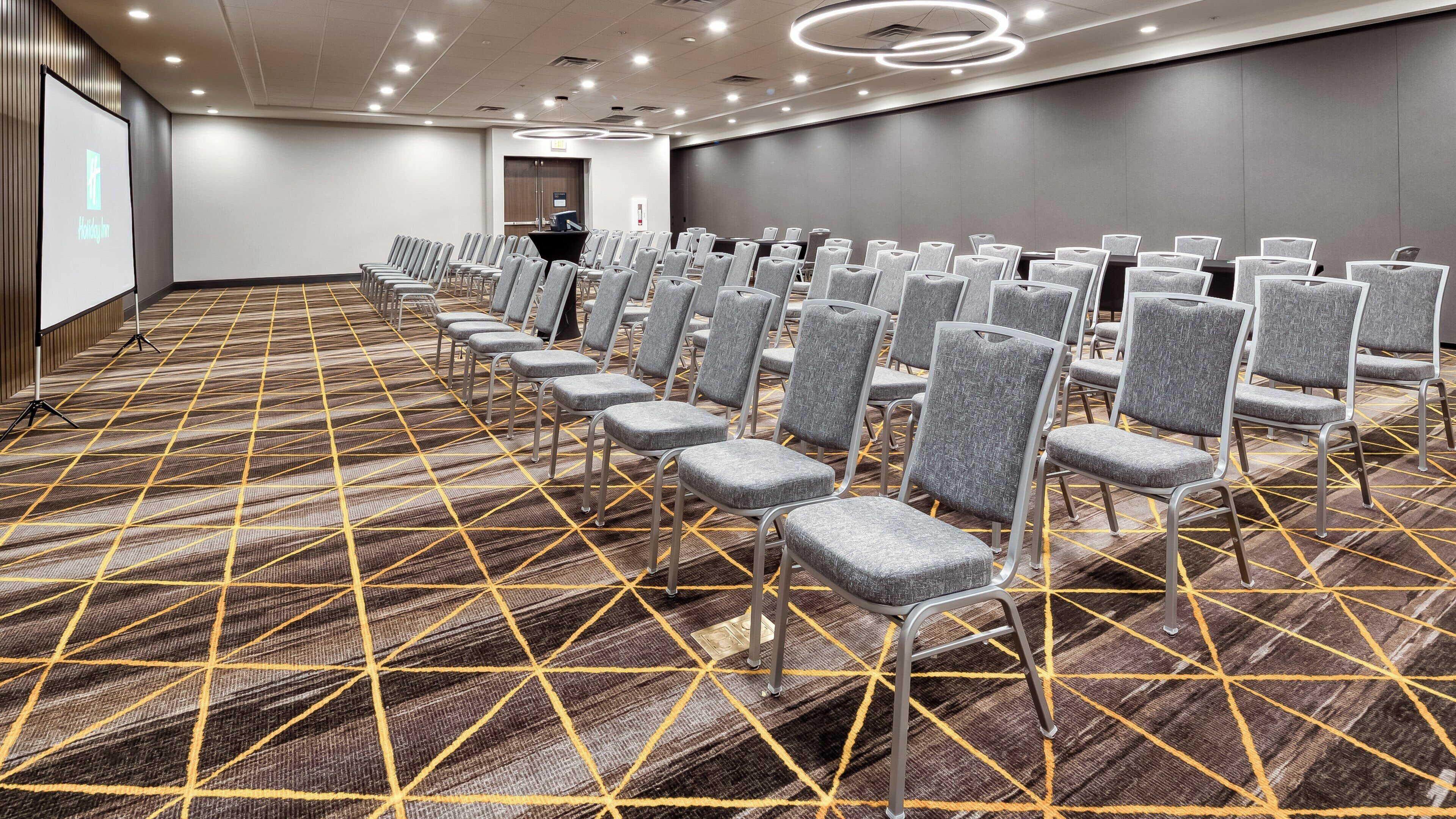 Une salle de conférence avec des rangées de chaises grises, une moquette à motifs et un écran de projection sur le côté gauche.
