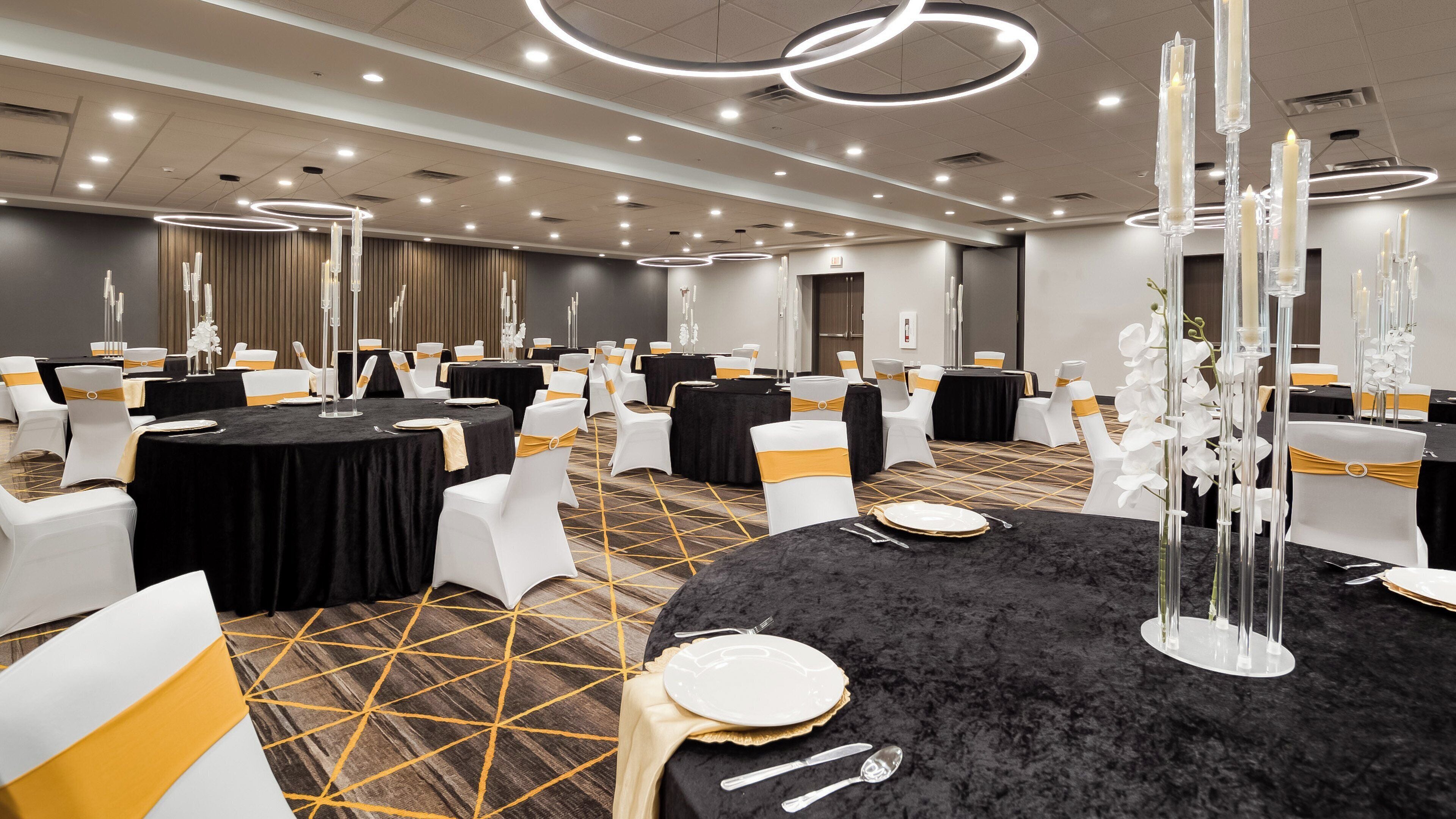 Une salle de conférence avec des tables rondes, des chaises blanches ornées de rubans jaunes, des nappes noires et de grands luminaires en forme de bougies.