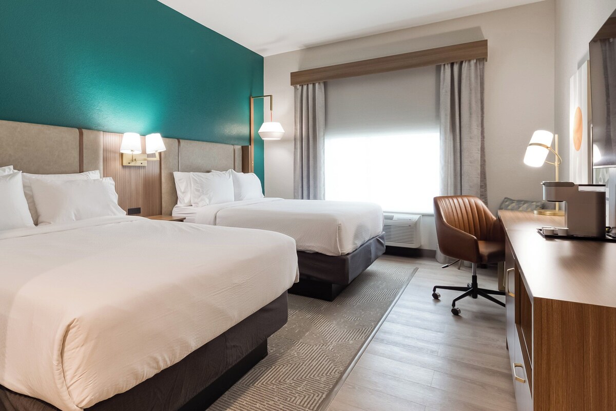Une chambre d’hôtel avec deux lits, un bureau, une chaise et une grande fenêtre avec des rideaux.