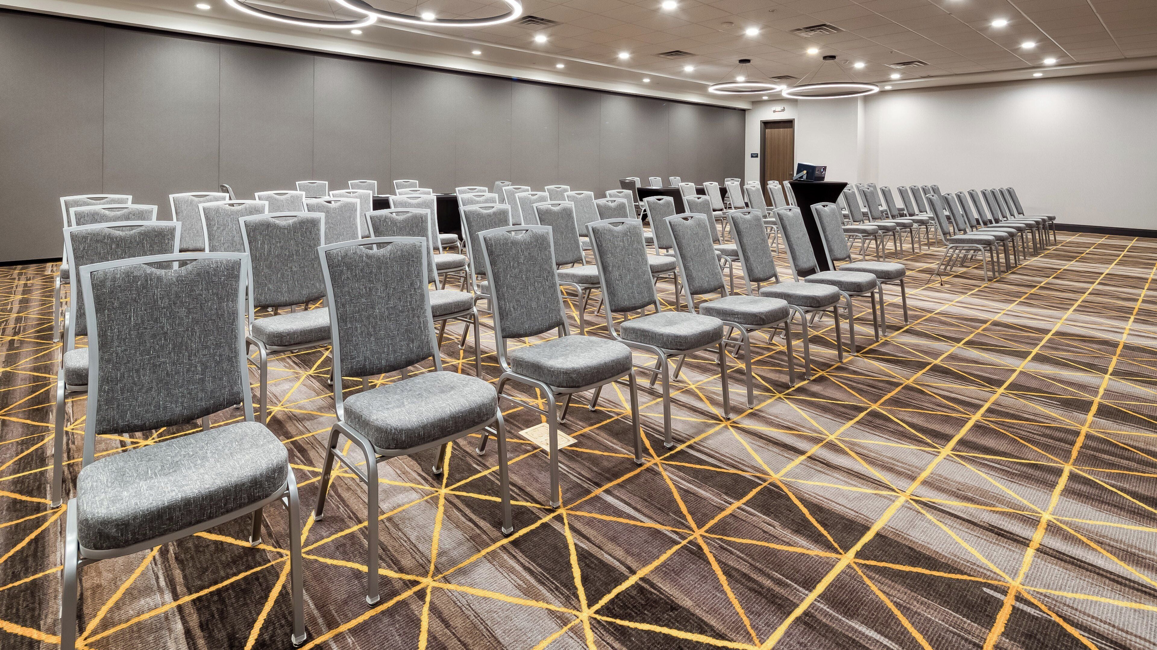 A conference room with rows of grey chairs, a carpet with a geometric pattern, and a modern ceiling with circular lighting fixtures.