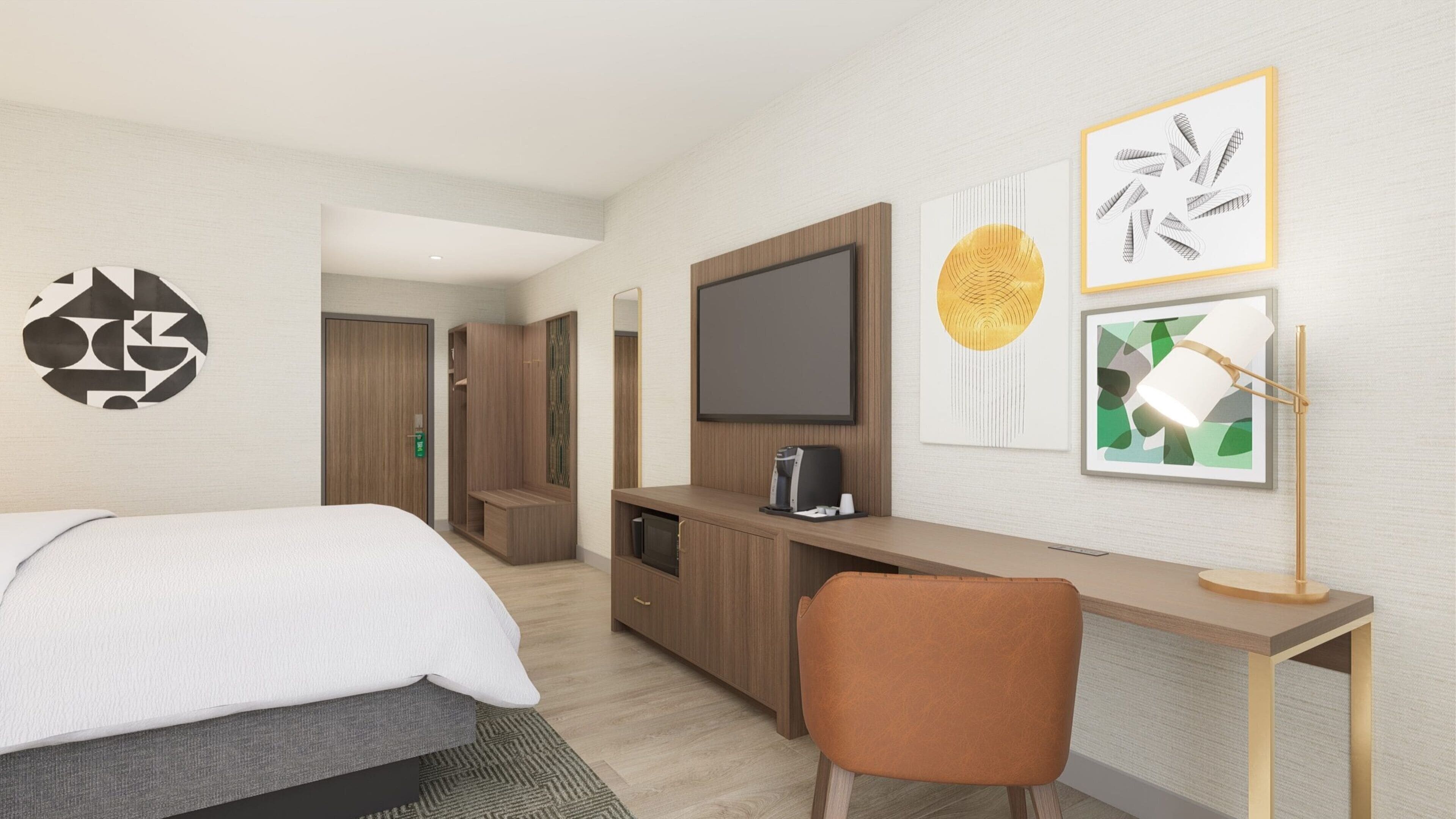 A modern hotel room with a bed, a desk with a computer, a TV, and artwork on the walls.
