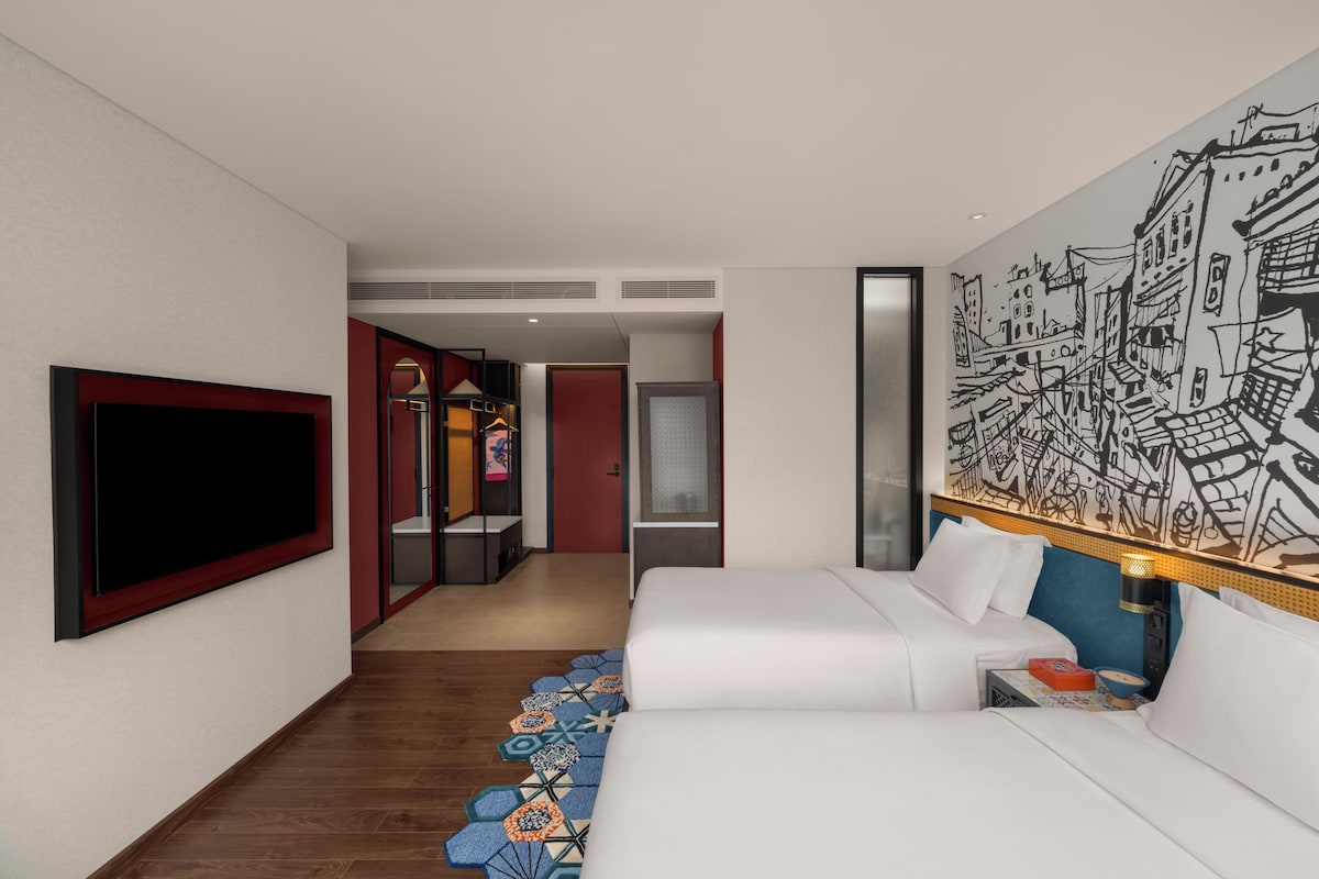 A modern hotel room with a large flat-screen TV, a bed with white linens, a colorful patterned rug, and a large abstract wall mural.