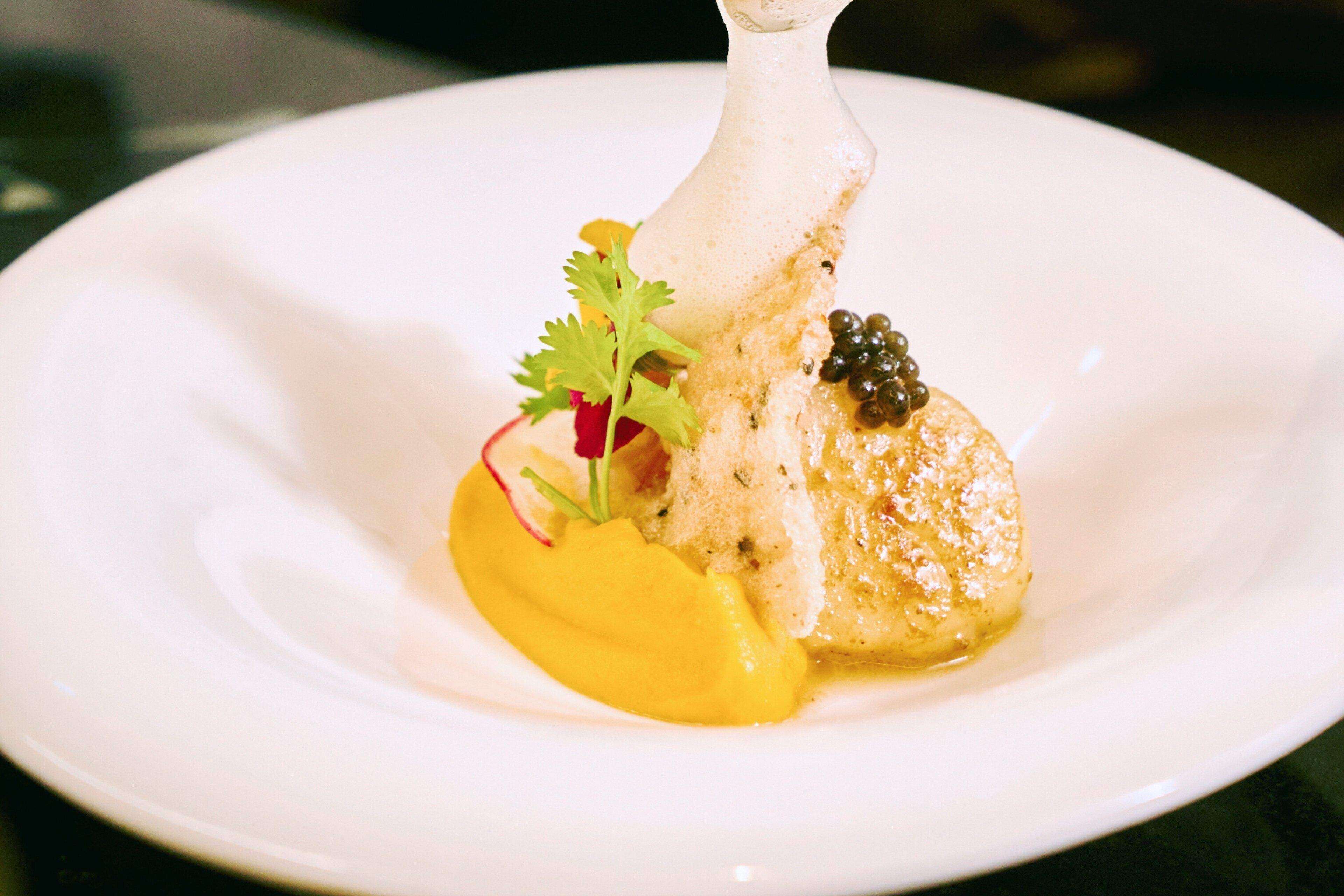 Une assiette blanche avec un morceau de poisson, une sauce jaune et quelques accompagnements.