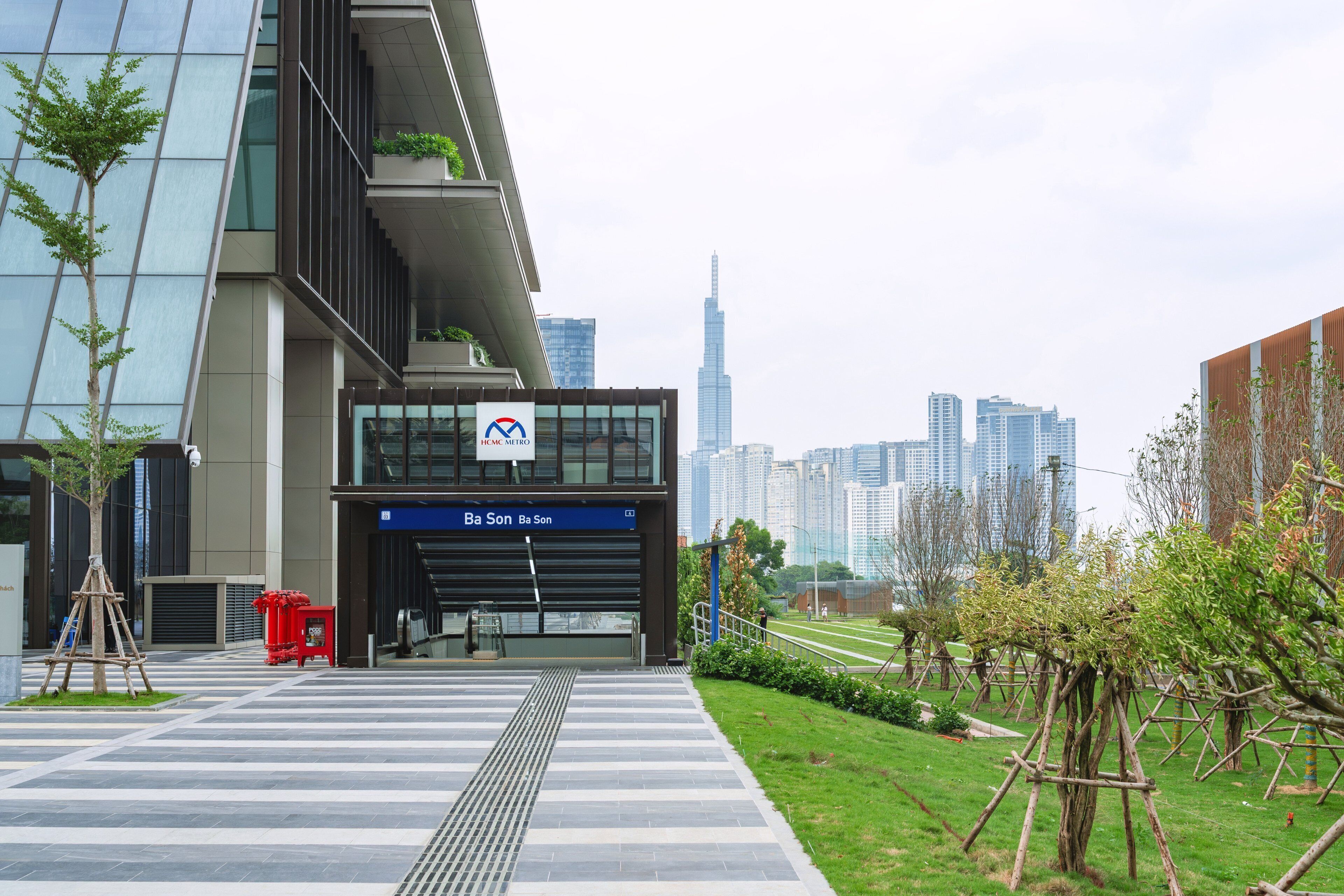 A modern building with a sign for HCMC Metro, a pedestrian walkway, and a city skyline in the background.