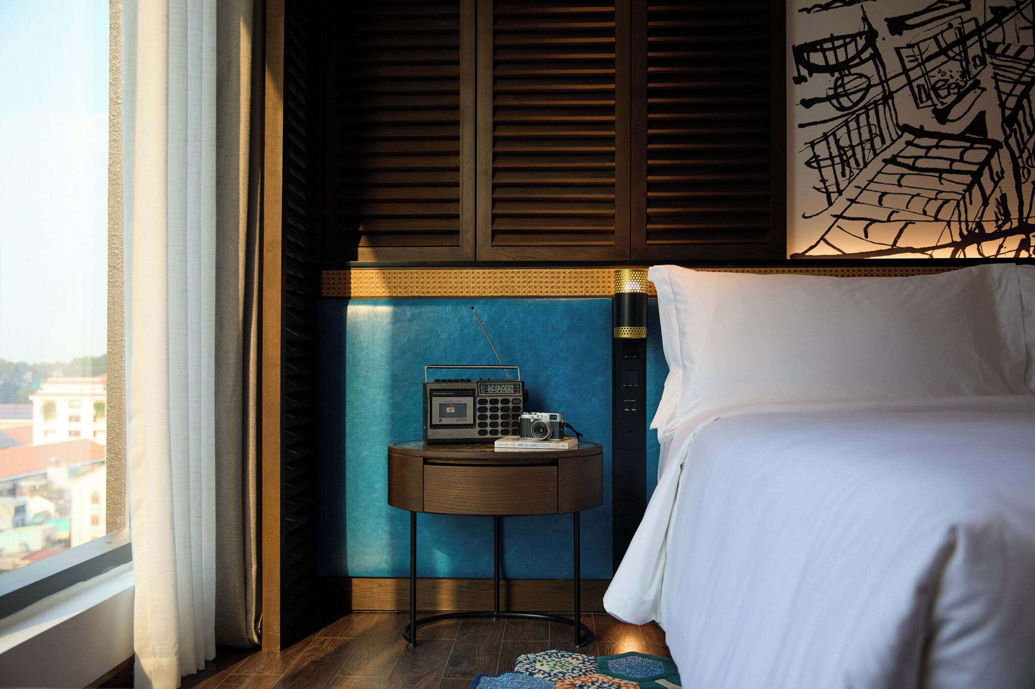 A hotel room with a bed, a small round table with a radio and camera, and a view of the city through the window.