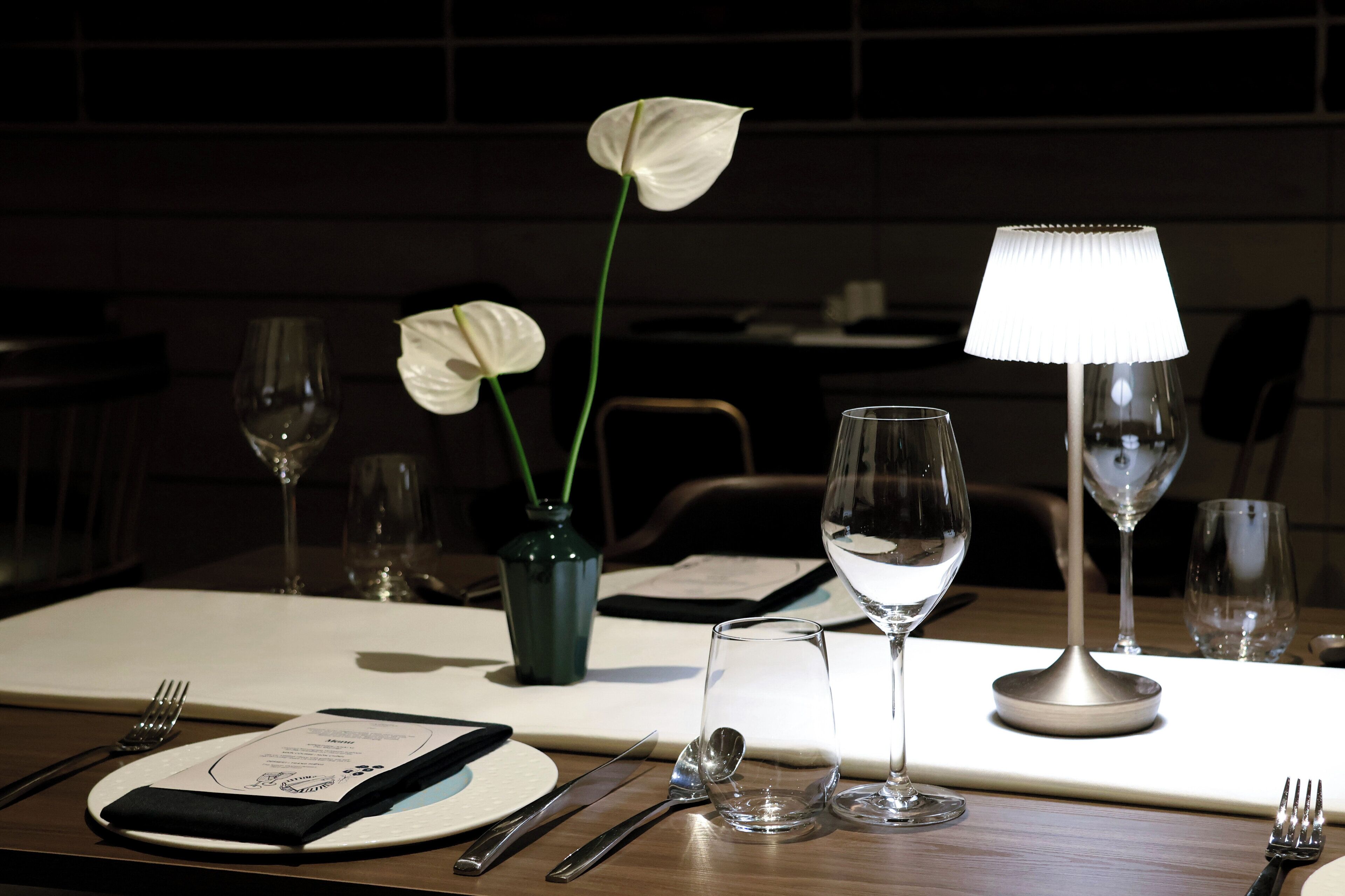 Ein Esstisch, gedeckt mit einer Vase weißer Blumen, einem Weinglas, einer Lampe und einer Speisekarte.
