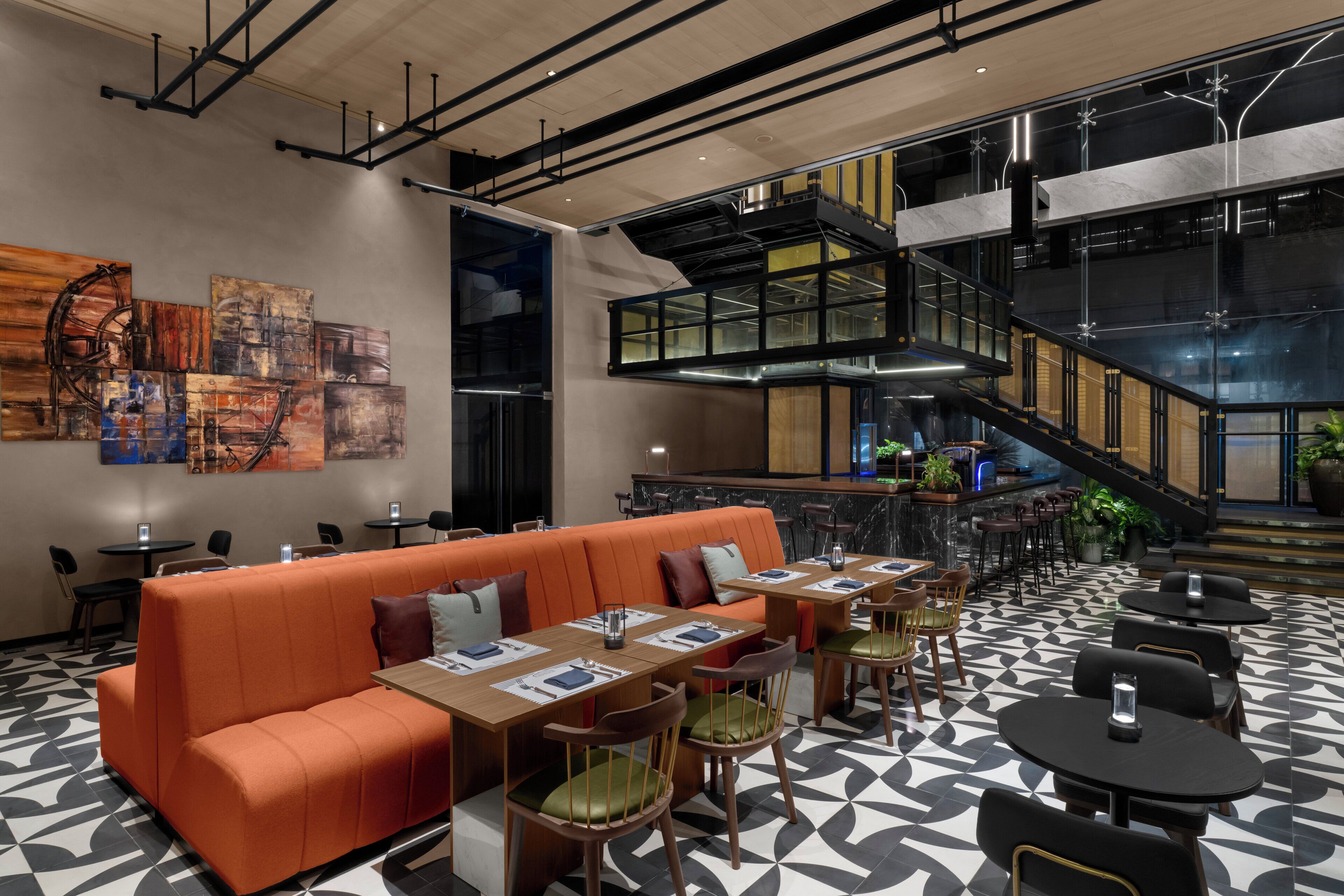 A modern interior with a central orange sofa, dining tables, and chairs, a staircase, and abstract art on the walls.
