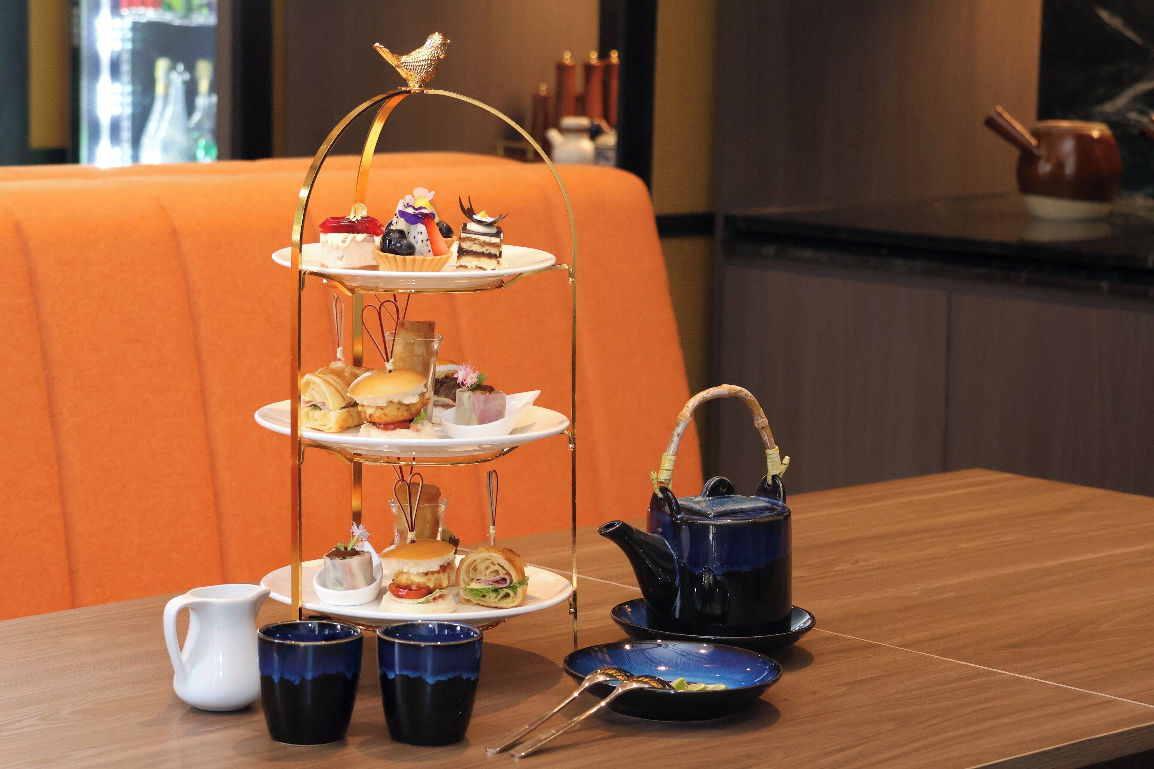 A three-tiered tea service set on a wooden table with a teapot and cups, accompanied by sandwiches and pastries, in a room with orange seating.