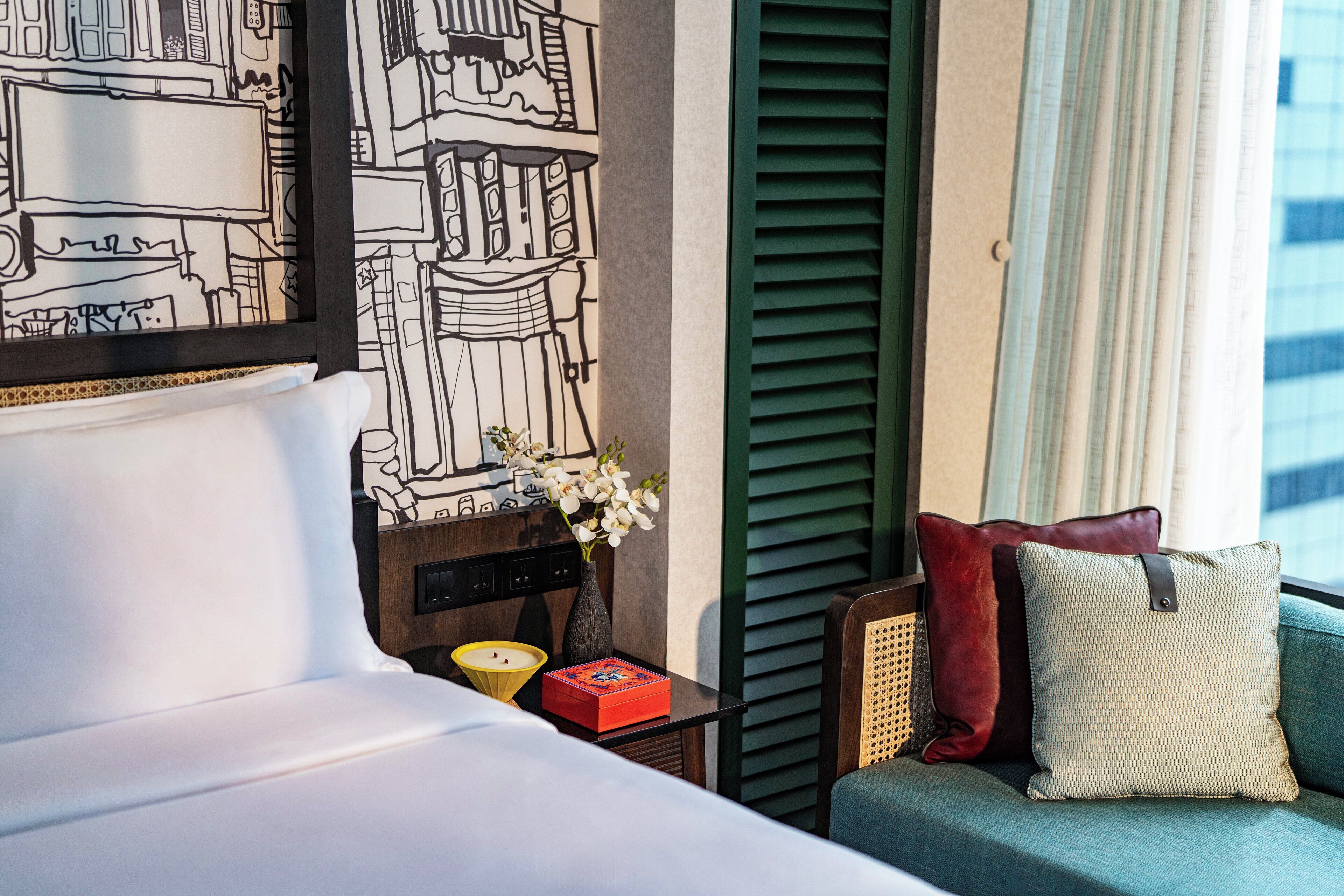 Een hotelkamer met een bed, een nachtkastje met een bloevas, een rood en groene bank en een muur met een zwart-wit stadsgezicht muurschildering.