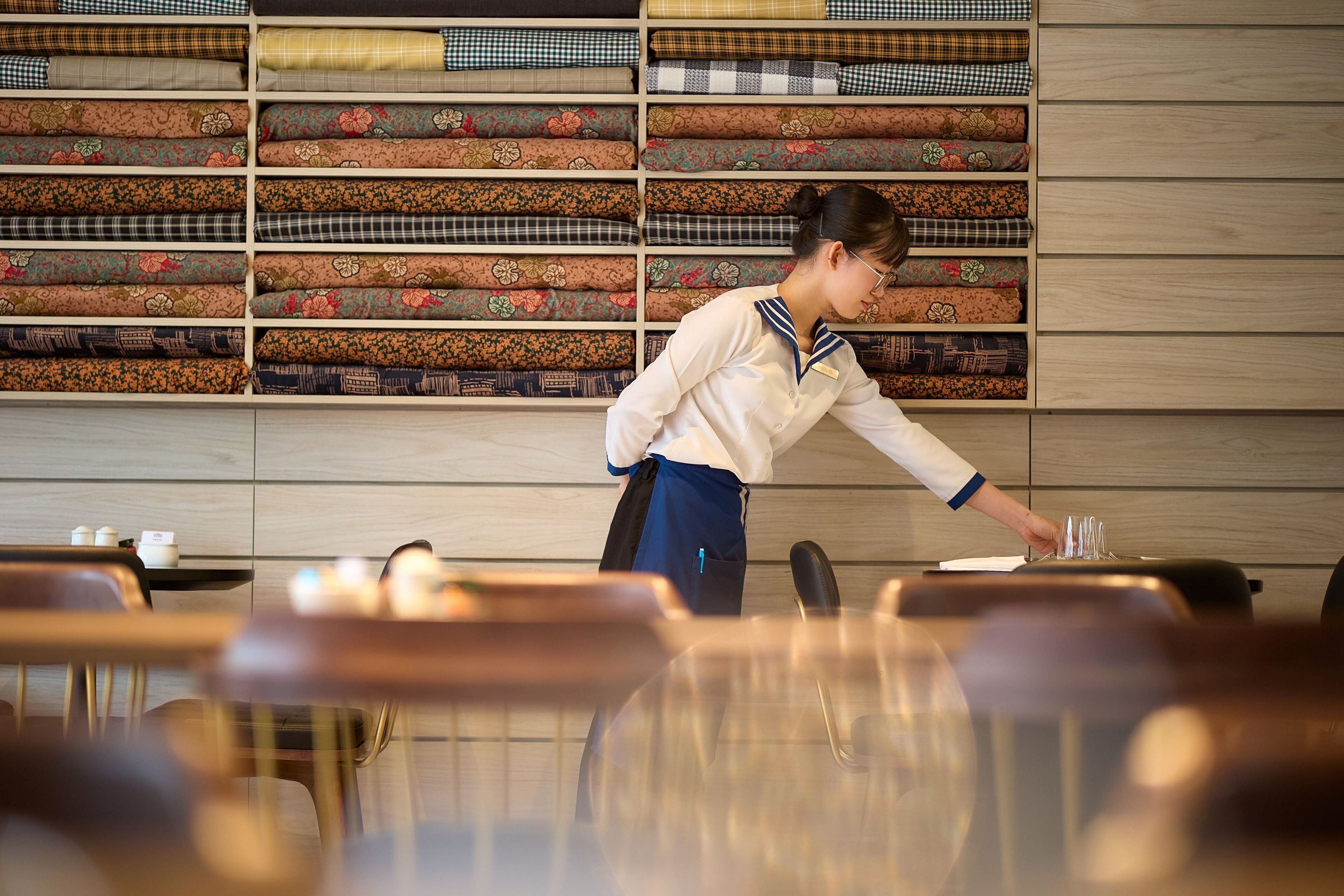 A person arranging items on a table in a room with a wooden wall and shelves of folded fabrics.