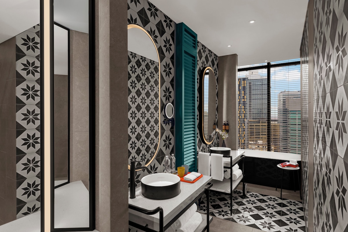 A modern bathroom with a large mirror, a round sink, and a window with blinds.