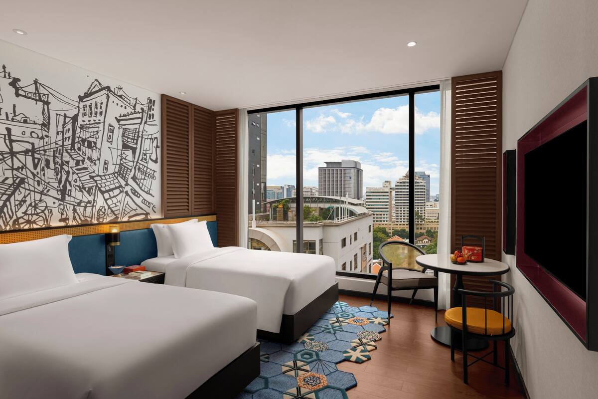 Une chambre d’hôtel avec deux lits, une grande fenêtre donnant sur la ville, une petite table avec une chaise et un téléviseur fixé au mur.