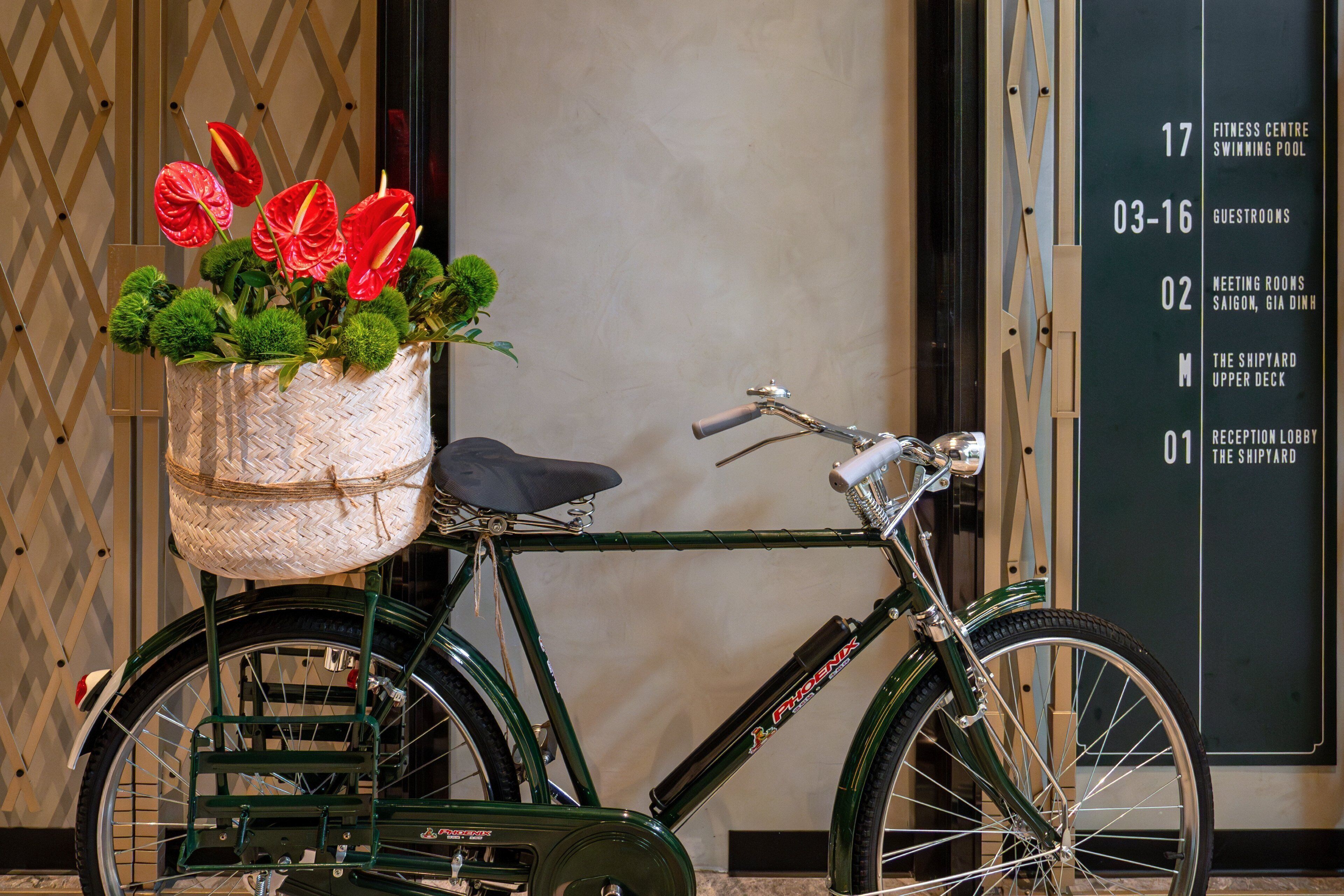 A green bicycle with a basket containing red flowers and green foliage, parked in front of a building with a wall-mounted sign listing room numbers and names.