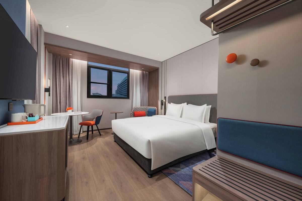 A modern hotel room with a large bed, a desk with a chair, a small table, and a window with curtains.