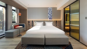 Une chambre d’hôtel moderne dotée d’un grand lit, d’un coin salon avec un banc et une petite table, ainsi que d’une grande fenêtre donnant sur l’extérieur.
