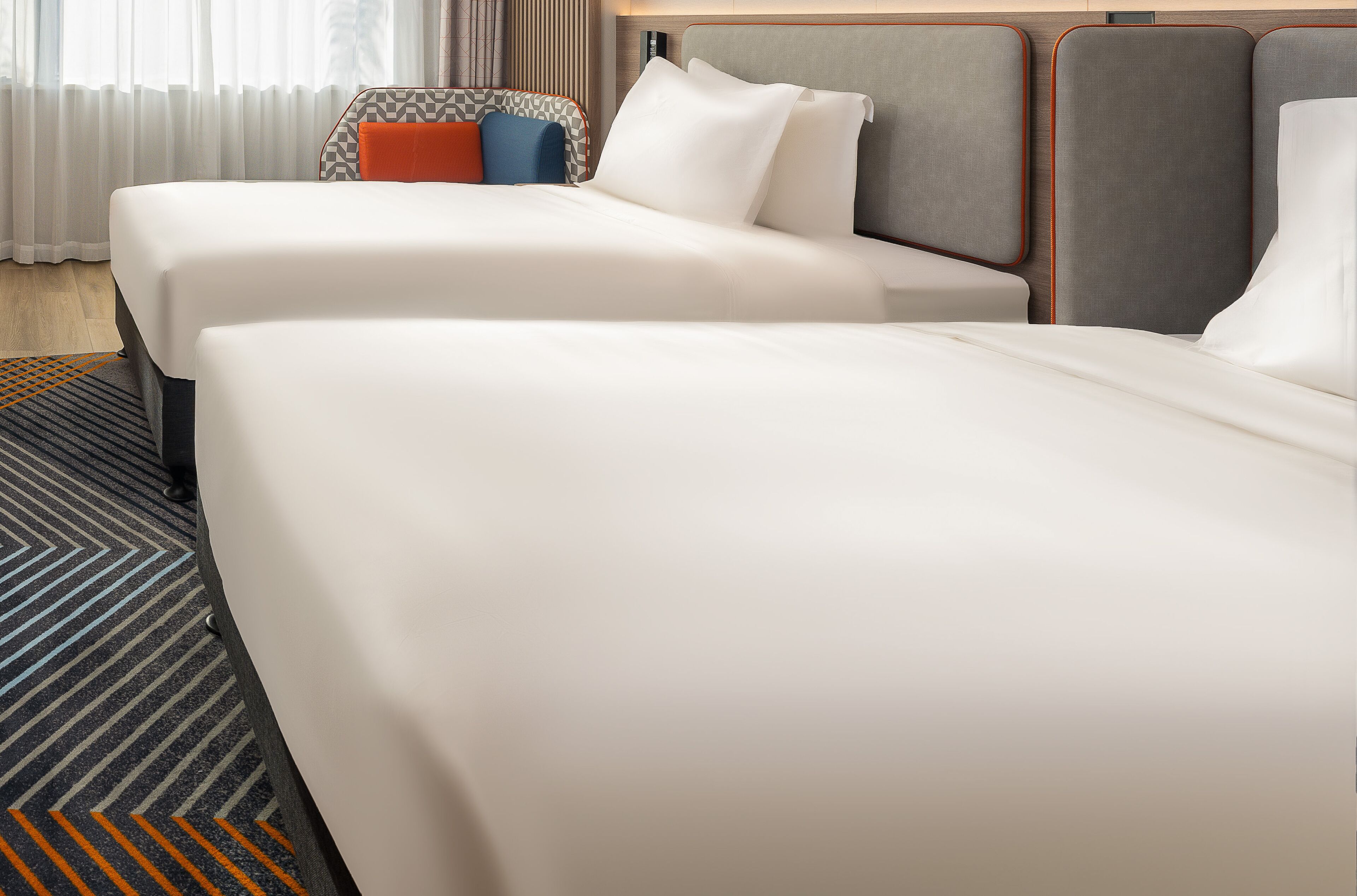 Habitación de hotel con dos camas, un sillón estampado y un ventanal con cortinas.