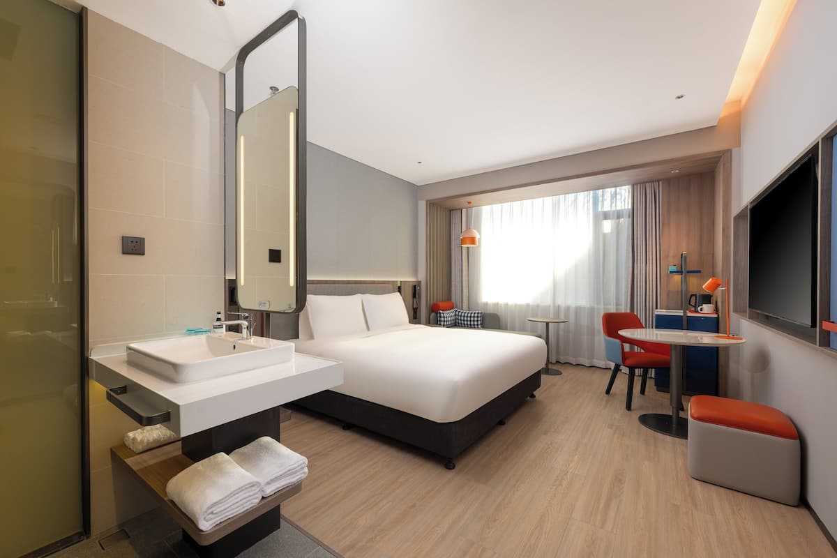 Ein modernes Hotelzimmer mit einem großen Bett, einem Badezimmer mit Waschbecken und Spiegel, einem Schreibtisch mit Stuhl und einem Fernseher.