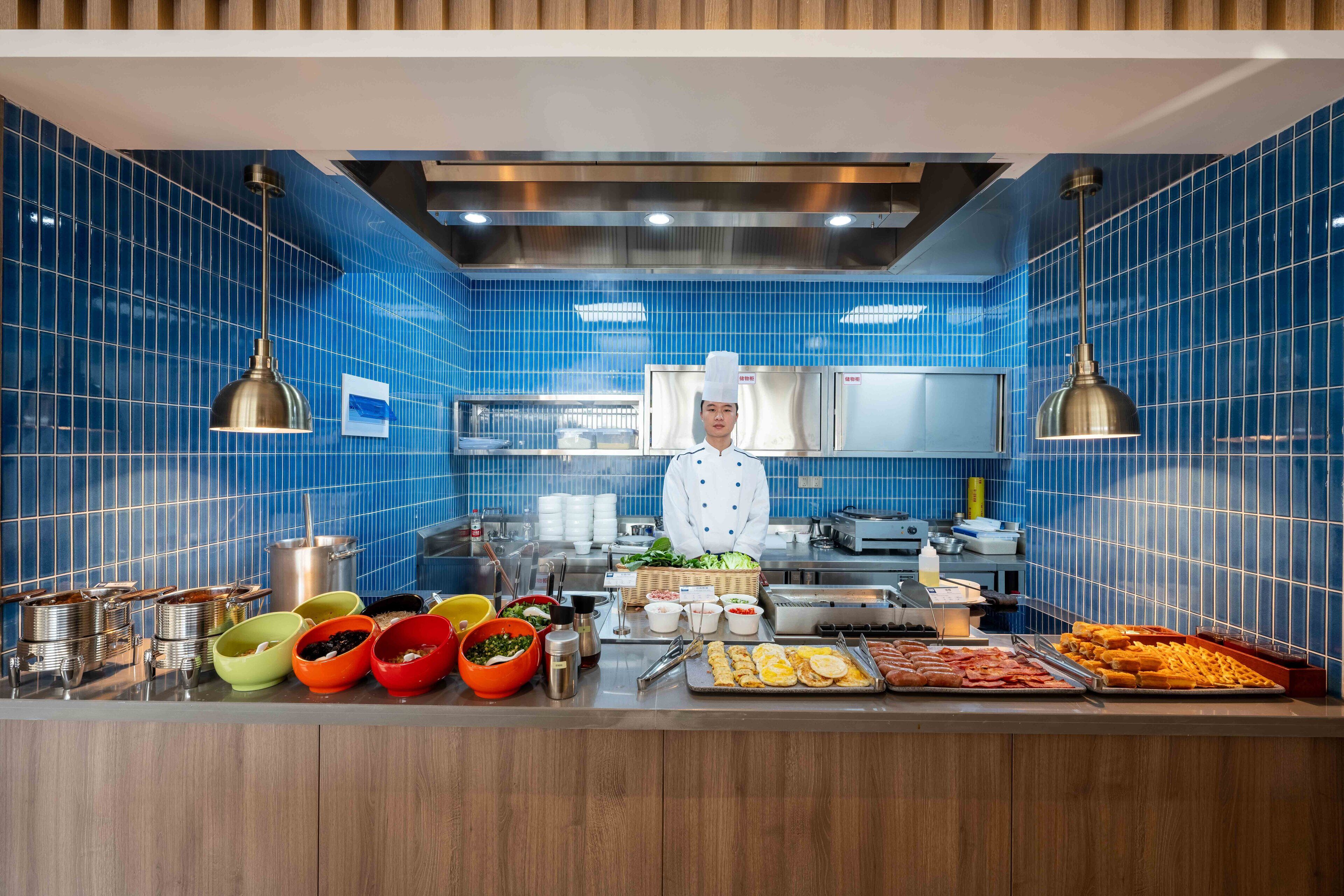A chef in a kitchen with blue tiled walls, stainless steel appliances, and a variety of food items displayed on the counter.