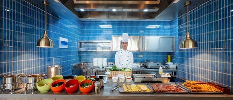 A chef in a kitchen with blue tiled walls, stainless steel appliances, and a variety of food items displayed on the counter.