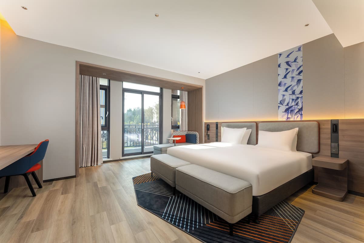 Une chambre d’hôtel moderne avec un grand lit, un bureau et une vue sur l’extérieur, visible à travers la porte vitrée.