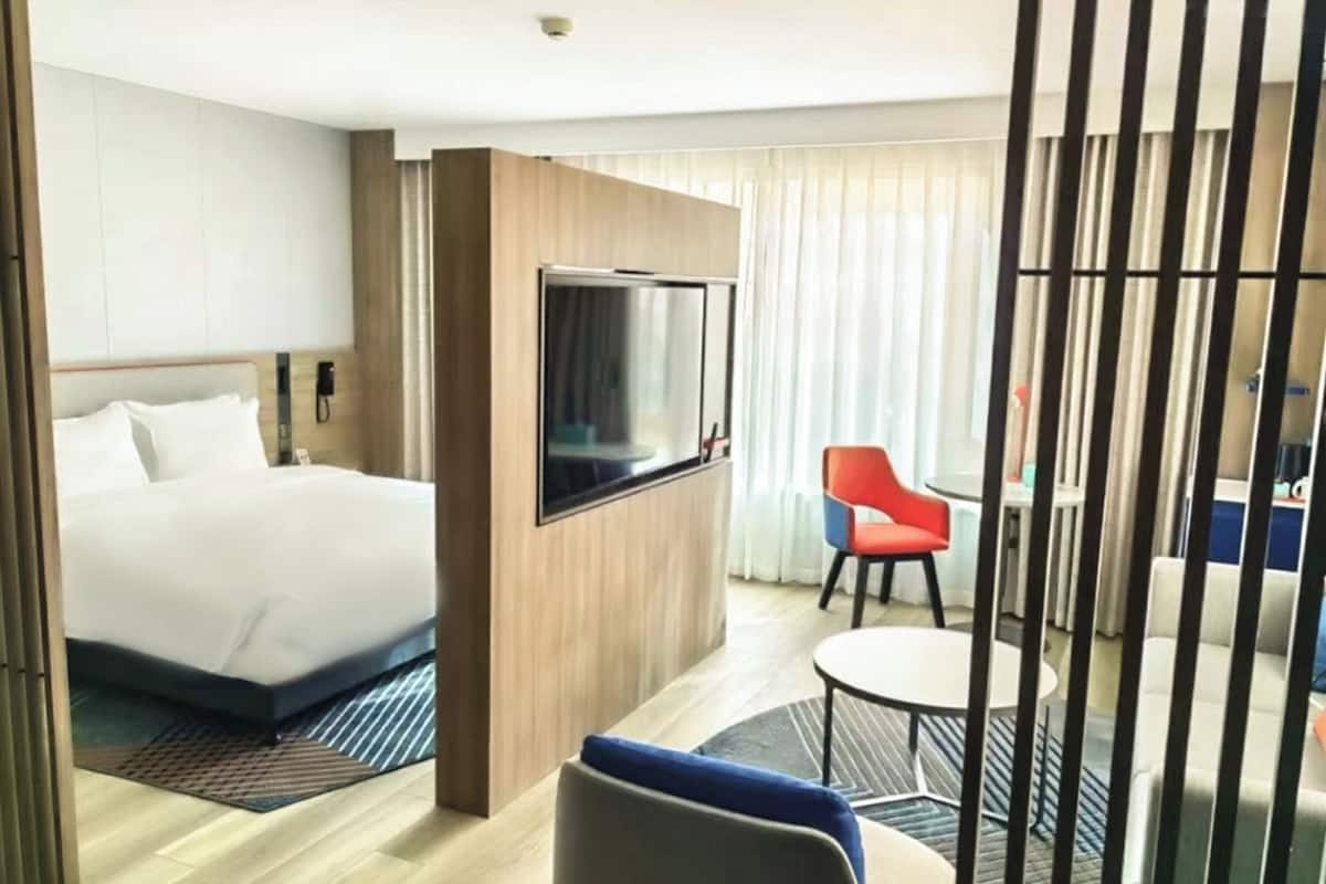 Ein Hotelzimmer mit einem Bett, einem an der Wand befestigten Fernseher, einem kleinen Tisch mit Stuhl und einem Fenster mit Vorhängen.
