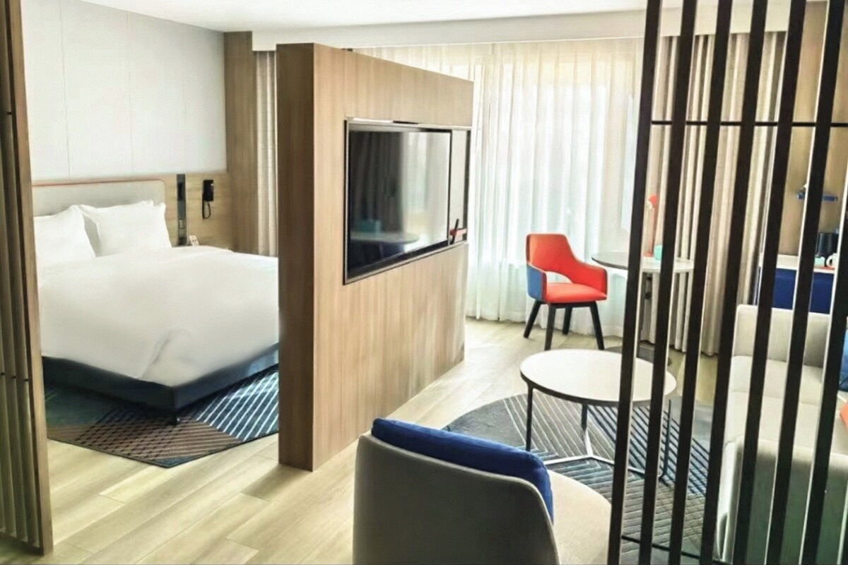 Une chambre d’hôtel avec un lit, un téléviseur fixé au mur, une petite table avec une chaise et une fenêtre avec des rideaux.