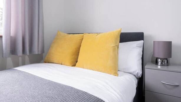 A bed with two pillows, a bedside table with a lamp, and a window with curtains. - 3 Bedroom Home With 2 Bathrooms & Free Parking (Bedford)