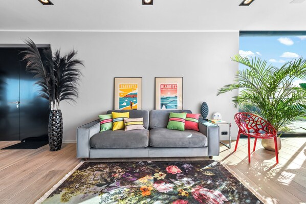 Un salon moderne avec un canapé gris, des coussins colorés, deux affiches encadrées, un grand vase avec des plumes noires, un fauteuil rouge et une grande plante en pot.