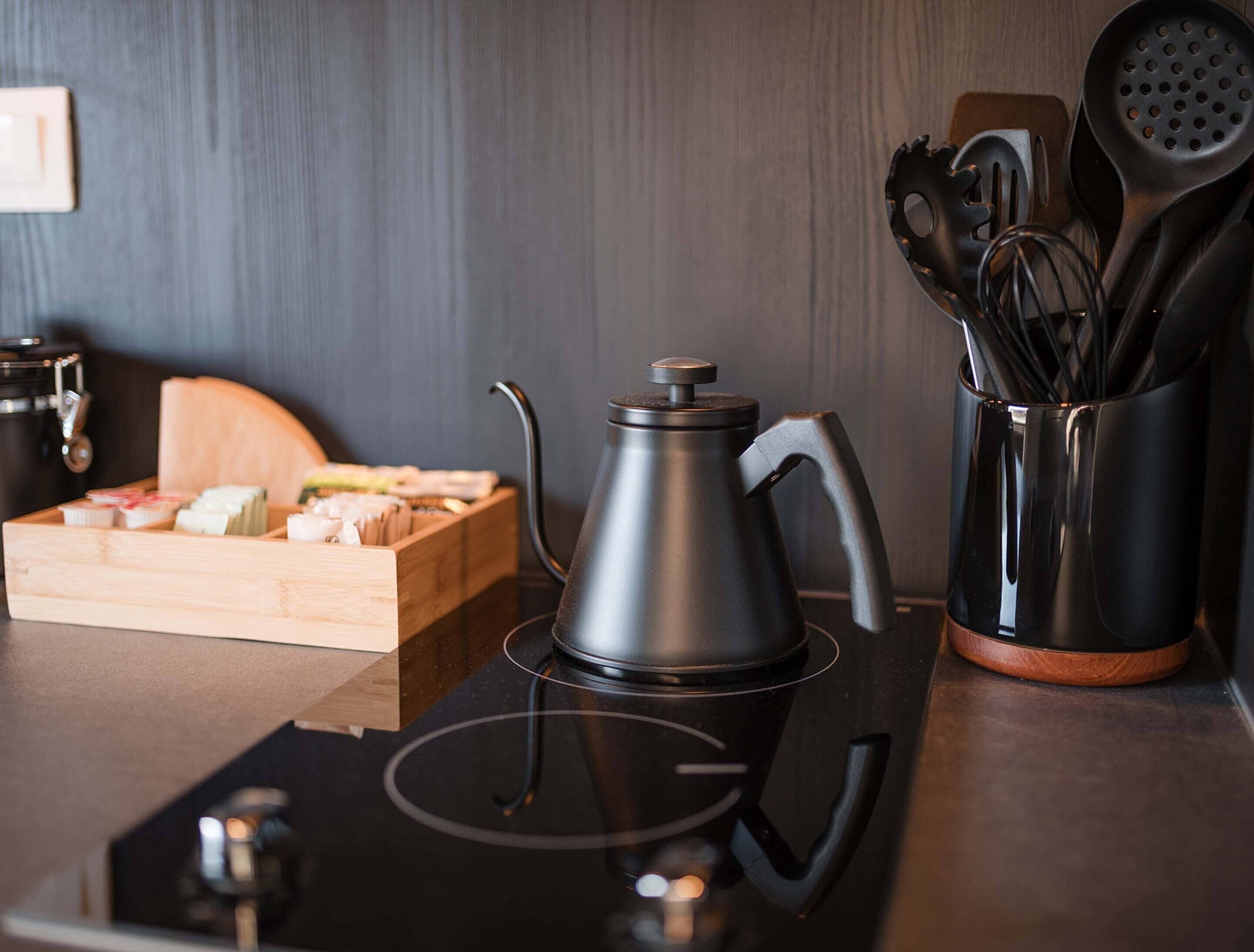 Una cocina con una tetera eléctrica negra, una tabla de madera con alimentos y un utensilio negro que contiene cucharas y un batidor.