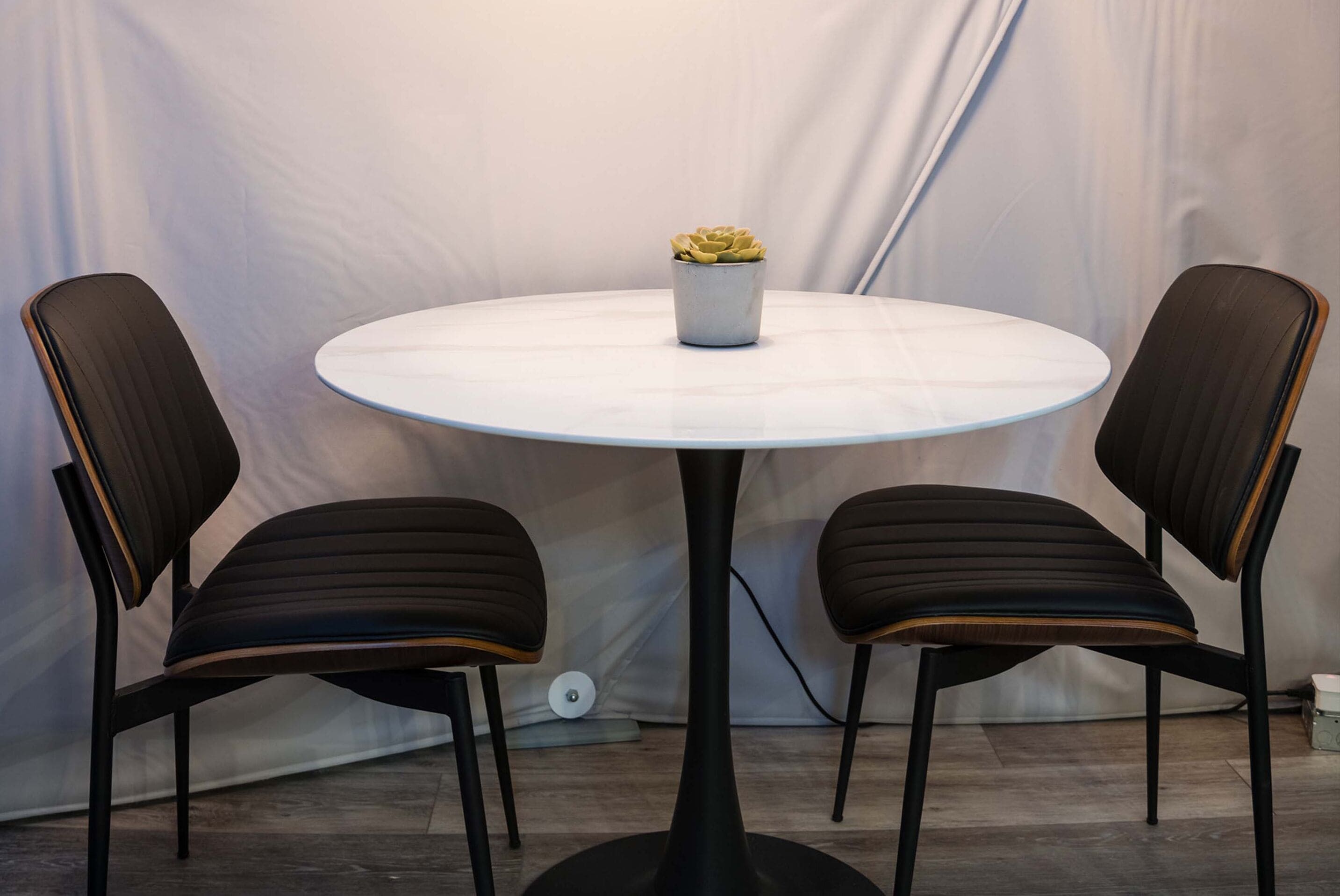 A round table with a white top and a single potted plant in the center. Two black chairs with wooden accents are positioned on either side of the table.