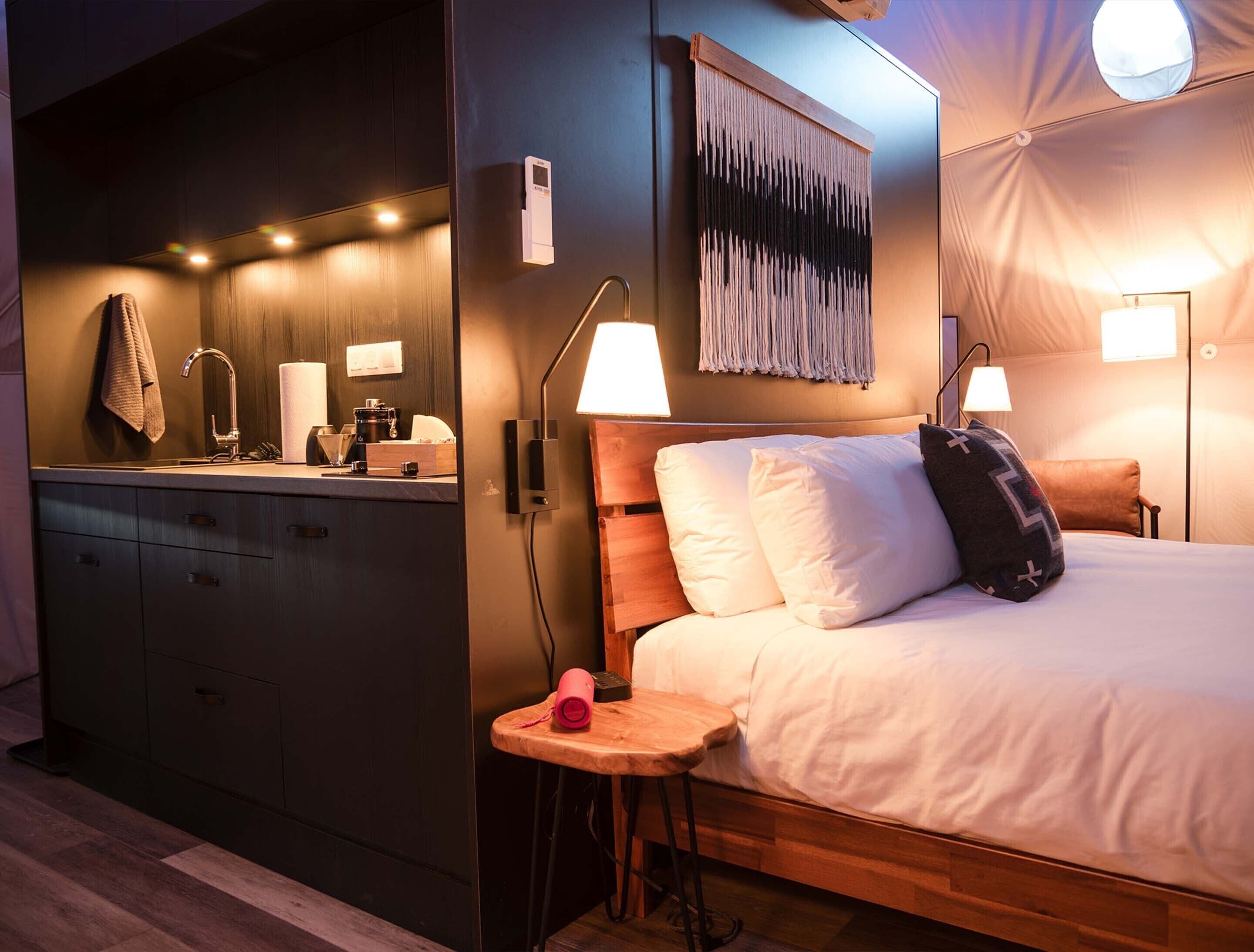 A modern hotel room with a wooden bed, bedside lamps, a small wooden stool, a mirror, and a kitchenette area.