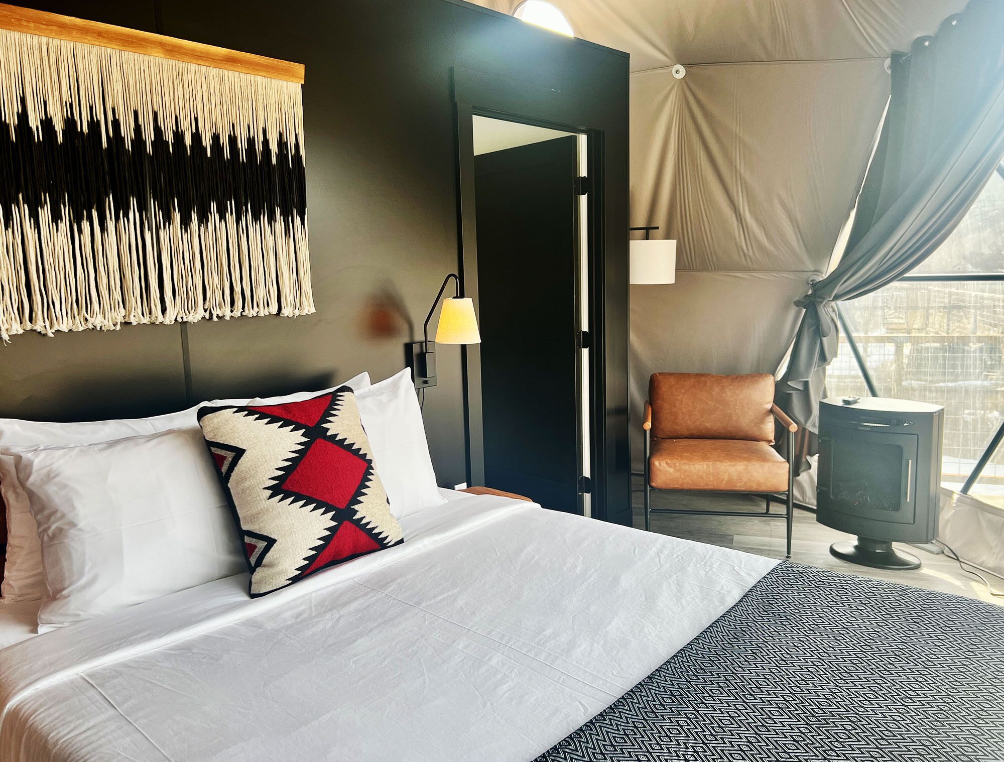 Una cama con almohadas blancas y estampadas, una lámpara, una silla y un calentador en una habitación con pared oscura y ventana con cortinas.
