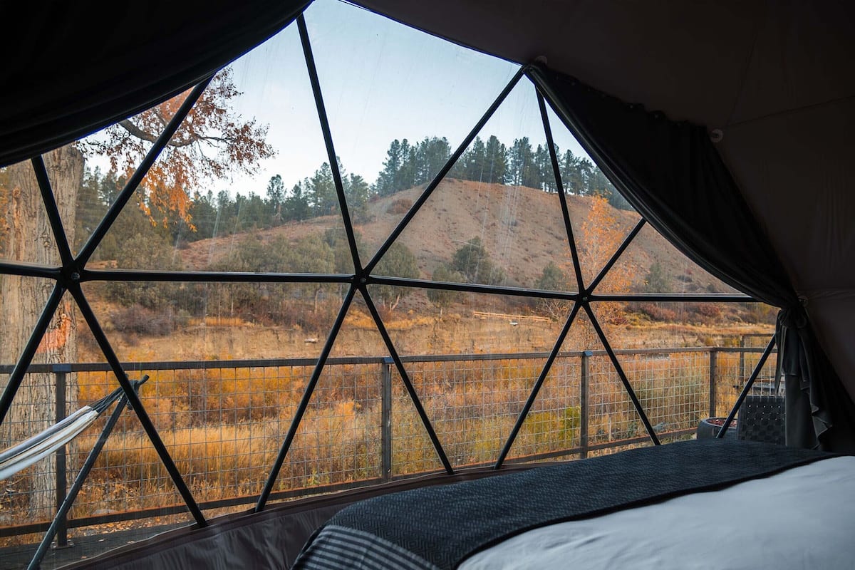 A tent with a geometric window design, a bed, and a view of a hilly landscape.