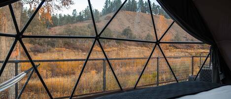 A tent with a geometric window design, a bed, and a view of a hilly landscape.
