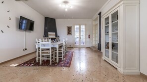 A dining area with a white table and chairs, a black wall-mounted TV, a patterned rug, and a cabinet with glass doors.