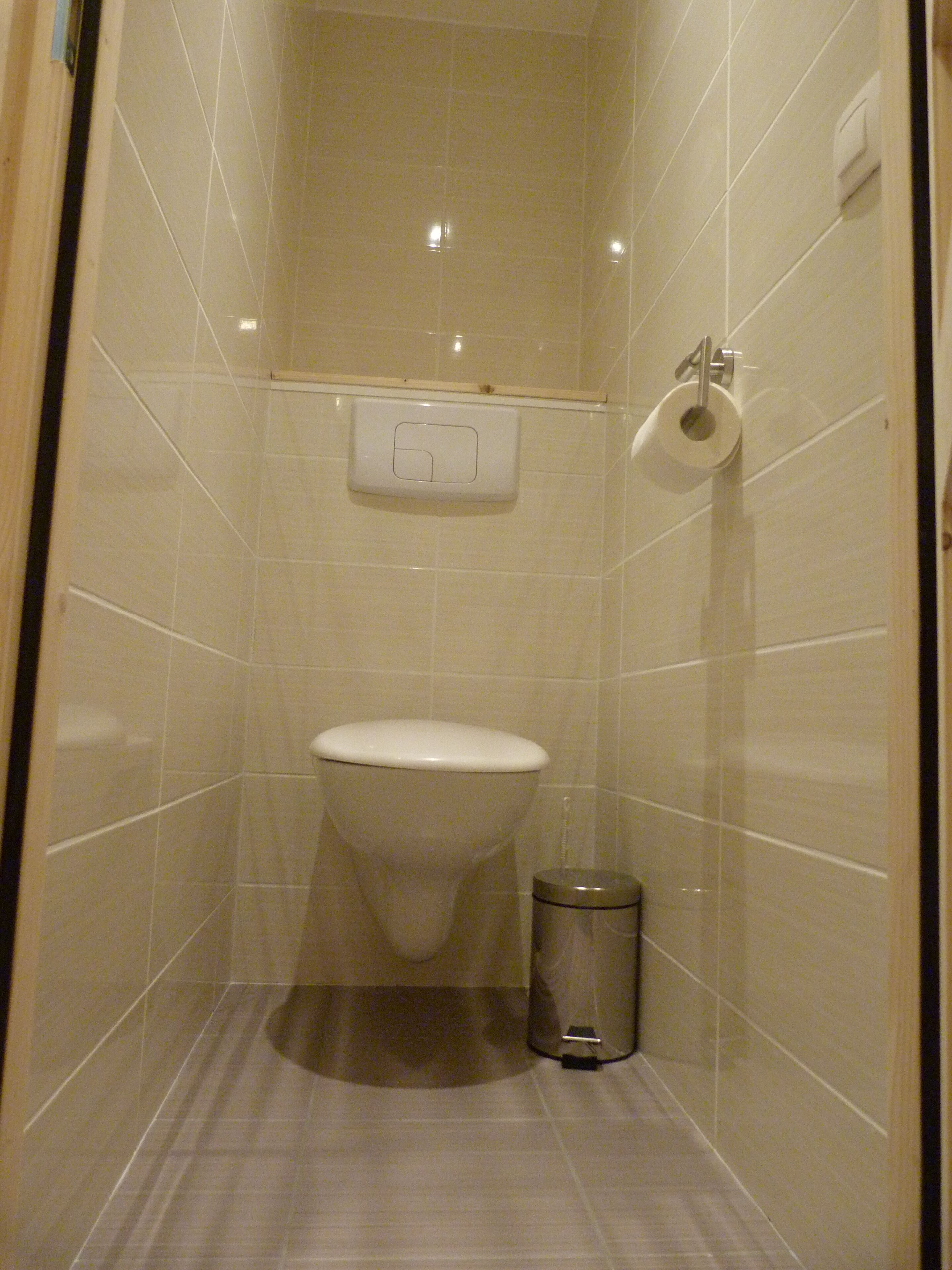 A small, tiled bathroom with a toilet, a trash can, and a paper towel dispenser.