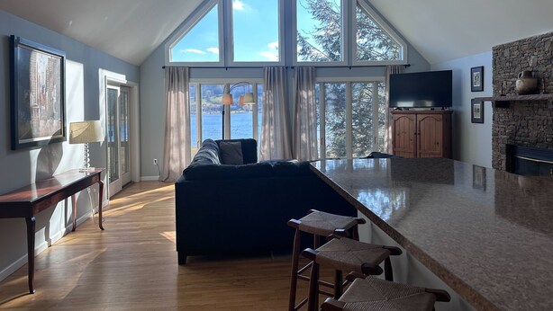 A spacious loft with a high ceiling, large windows offering a view of the outdoors, a cozy seating area, and a kitchen with a stone accent wall.