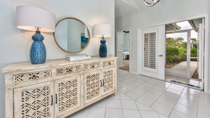 Um corredor com piso de azulejos brancos, um armário de madeira com entalhes elaborados e um espelho grande. Há uma porta que dá para uma varanda com vista para o verde.