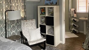A bedroom with a bed, a chair, a bookshelf, a ceiling fan, and a bathroom visible through an open door.